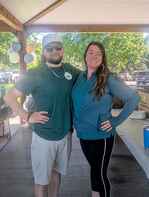 Tara and Anthony stand in a covered outdoor space and smile. They each have an arm around each other and are both wearing Heritage NW Group shirts.