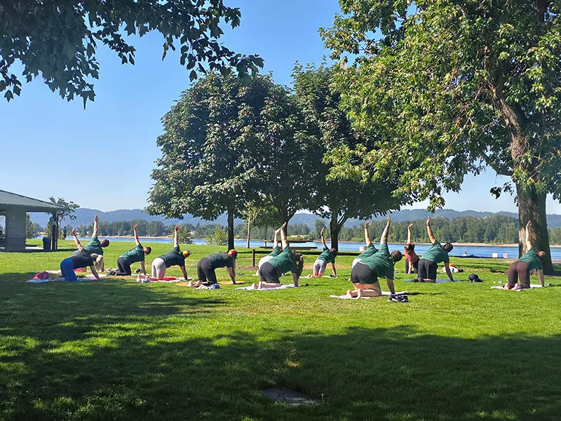 Heritage employees do yoga in the grass at the park. In the background trees and water shine in the sun. Everyone is on a yoga mat on hands and knees with one arm stretched into the air.