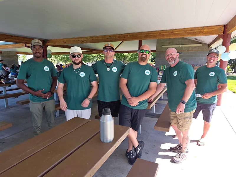 Six Heritage employees stand in a covered outdoor space between picnic tables. They are all wearing matching Heritage NW Group green shirts.