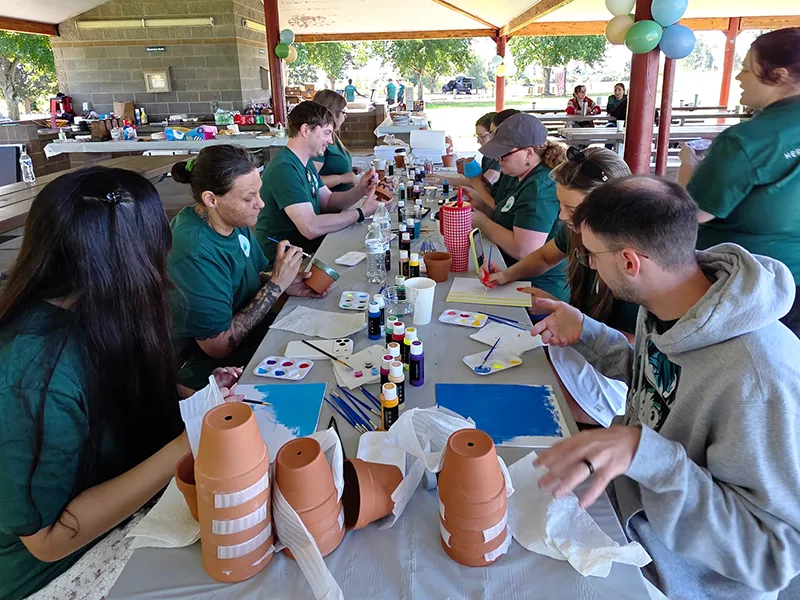 Heritage employees sit at a covered picnic table in a covered outdoor space and work on individual painting projects. Some people are painting terra cotta plant pots. Some people paint canvas or paper.