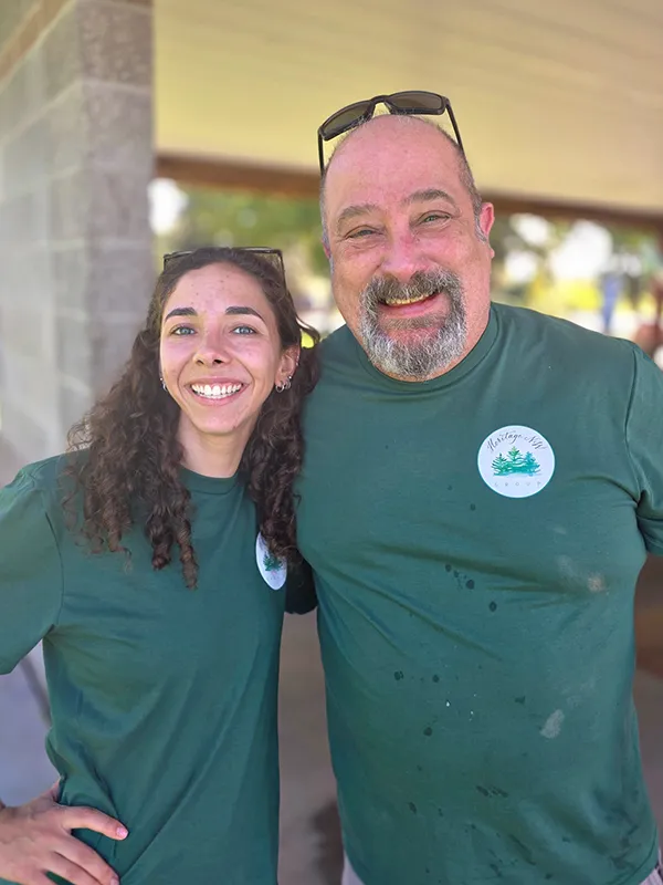 Two Heritage employees in green Heritage NW Group shirts smile with an arm around each other.