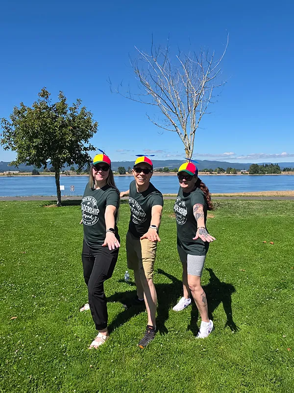 Three Heritage employees stand in a small lunge with one arm stretched forward and the other arm stretched backwards. They are all wearing multicolored red, yellow, and green ball caps with propelers on top and matching green shirts. The shirts are only partially visible but they appear to say We're that Leadership squad they told you about.