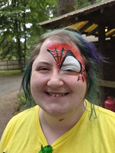 Person with colorful hair smiles at the camera with spiderman style webs face painted around one eye