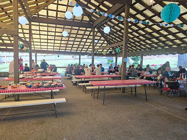 Covered pavilion with people sitting at picnic tables covered in food and red checked tableclothes.
