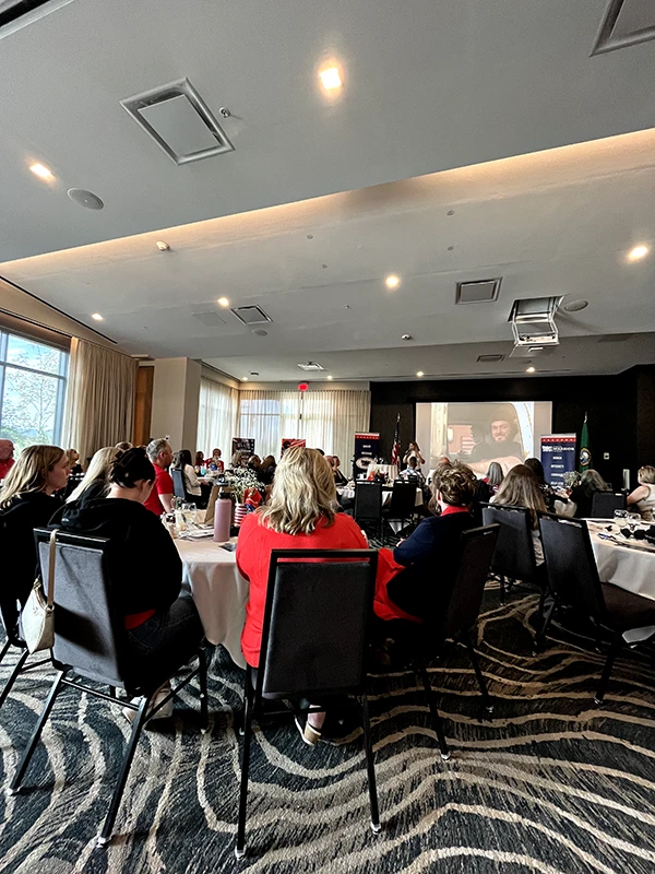 People sit at tables in a ballroom watching a presentation on a projector screen. AWI banners are on either side of the screen.