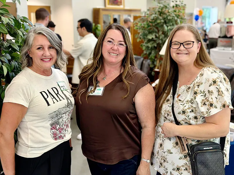 Three people, including Tara stand together and smile. They are wearing nametags and in the background you can see more people standing together and talking