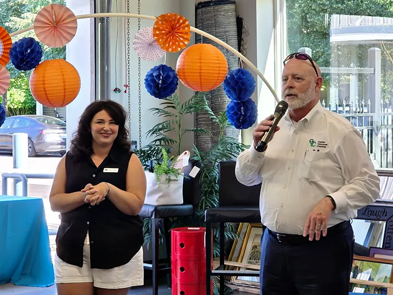 GVC employee stands in front of orange and blue paper ball decorations. He is speaking into a microphone and another person is standing nearby, smiling and clasping her hands comfortably in front of her body.