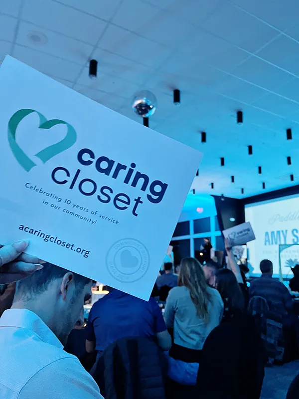 People sit in a room filled with blue lighting around tables. A hand is holding a paper that says caring closet Celebrating 10 years of service in our community