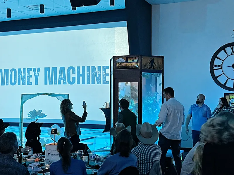 The money machine is running. It is a phonebooth style box with paper bills being blown around for participants to grab. A person is taking a photo of the money machine running with a cellphone camera while others watch