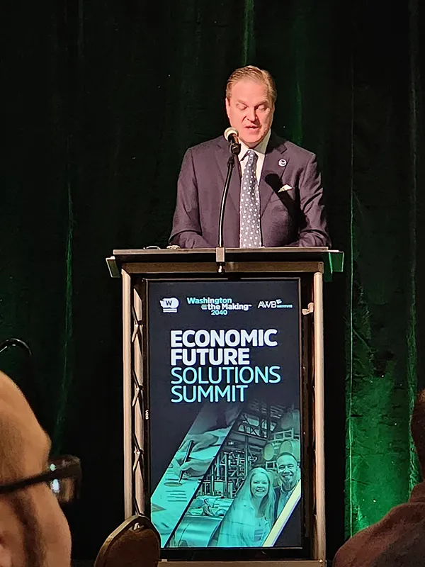 A man in a suit stands at a podium with a screen on the front with the words Economic Future Solutions Summit. There is a green curtain behind him. He is looking down at the podium and speaking into a microphone.