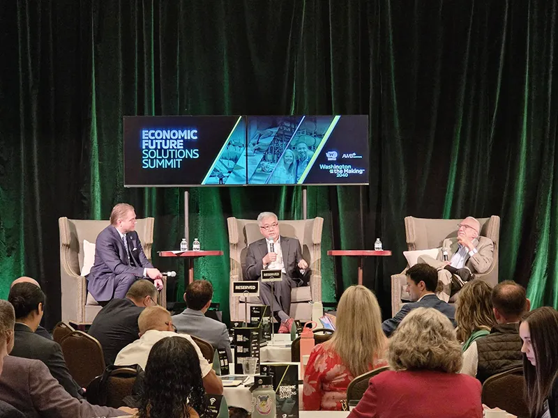 Three people sit in chairs on a stage in front of a green curtain and a screen displaying Economic Future Solutions Summit. People are sitting at tables in front of the stage listening to the speakers.