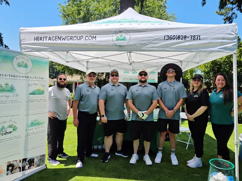 Heritage employees stand under a pop up shade in front of a table covered in green with Heritage NW group materials and branded gifts. There is a pull up banner sign that has information about the Heritage companies.
