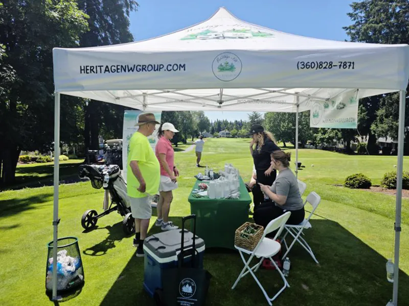 Heritage employees are seated and standing behind the green covered table talking to people. Golf is being played in the background. It is a sunny day and the grass is very green.