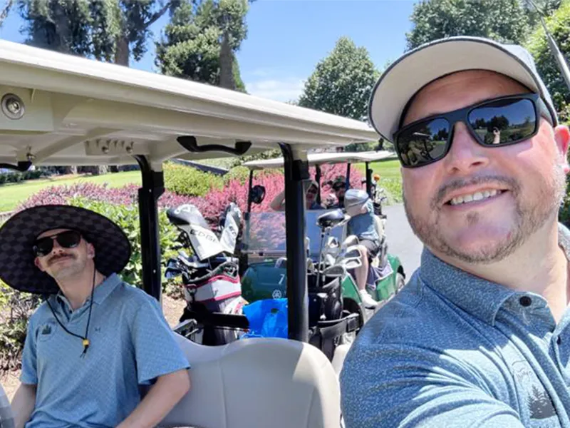 Heritage employees sit in a golf cart smiling and preparing to play. They are wearing hats and sunglasses and their golf clubs can be seen in the back of the golf cart. Another golf cart is in the background. There are flowering bushes and lots of green trees and grass.