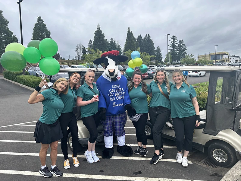 The team stands in front of golf carts holding gree balloons and posing with the Chick-fil-A cow.