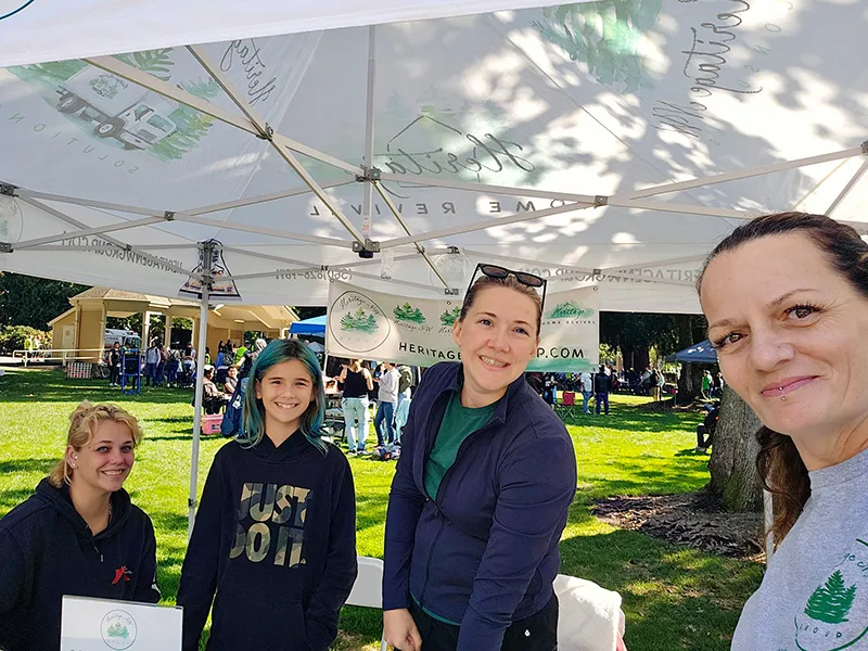 Heritage employees sit and stand around the table under the Heritage NW Group shade tent.