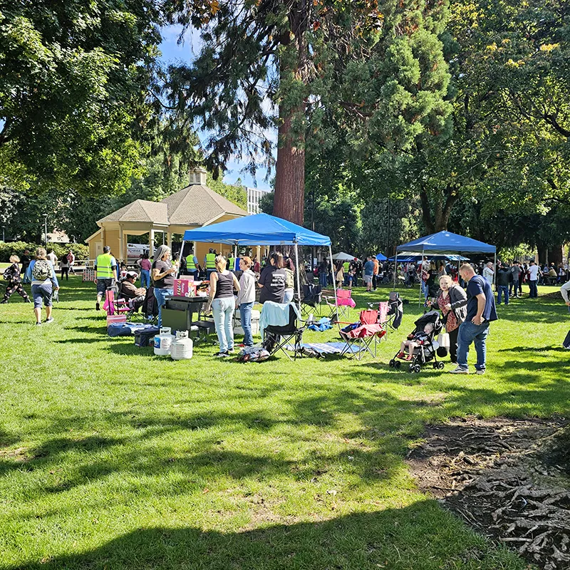 People gathered in gropus at the Hands Across the Bridge event. There are many pop up shades with tables set up, camping chairs and picnic blankets spread on the grass under the trees.