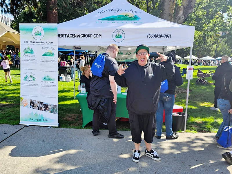 The Heritage NW Group pop up shade tent with people talking to Heritage employees. A person wearing black clothes and a ball cap is smiling and making the peace sign with both hands.