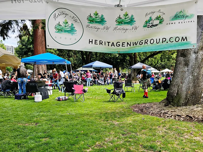 A view of the crowd of people gathered at the Hands Across the Bridge event. The picture is taken from underneath the Heritage NW Group shade tent with a banner displaying the Heritage brands and website heritagenwgroup.com.