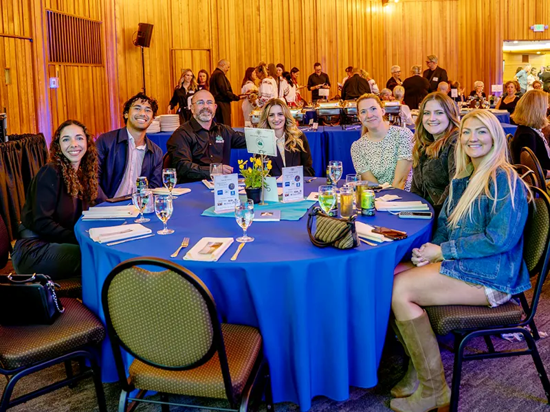 Seven Heritage NW Group employees sit at a round table with a blue tablecloth. Catering is visible in the background with warmers of food.