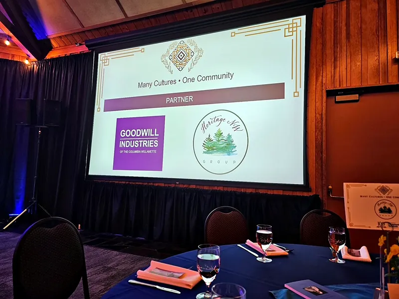 A projector screen displays a Partner slide at Many Cultures, One Community. Goodwill Industries and Heritage NW Group logos are displayed.