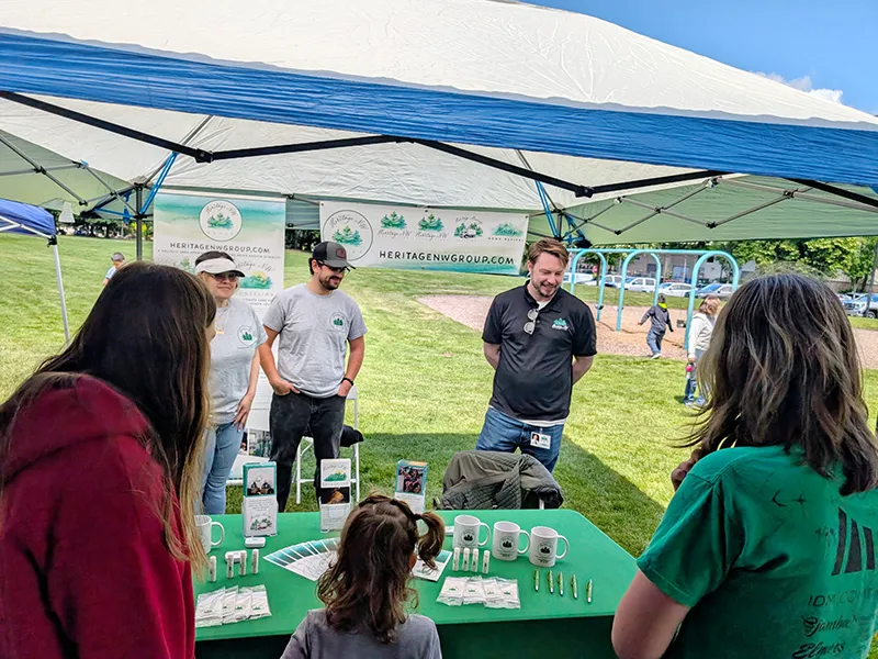 Heritage employees stand under a pop up shade at a table covered in green with Heritage NW group materials and branded gifts. There are people with their backs to the camera who are talking to the Heritage employees and looking at the items on the table.