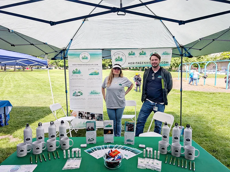 Heritage employees stand under a pop up shade at a table covered in green with Heritage NW group materials and branded gifts.