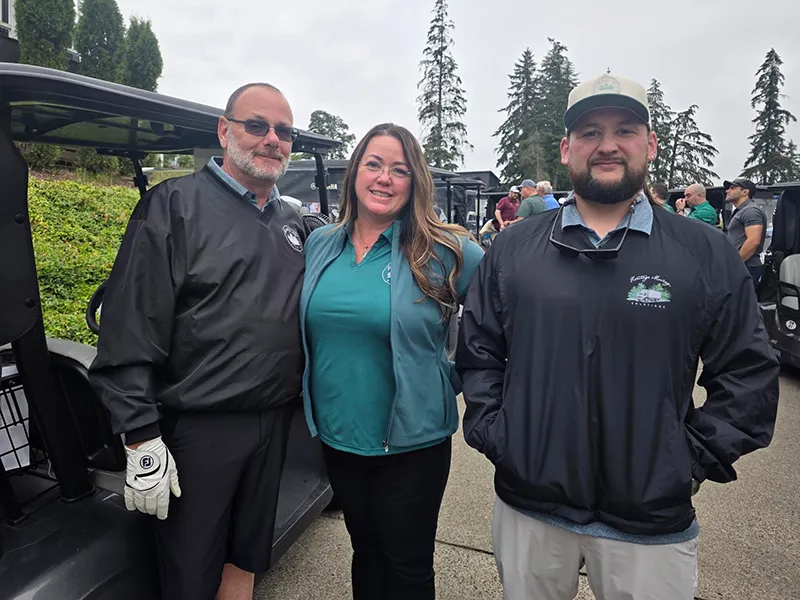 Jim, Tara, and Anthony stand beside a golf cart at the tournament.