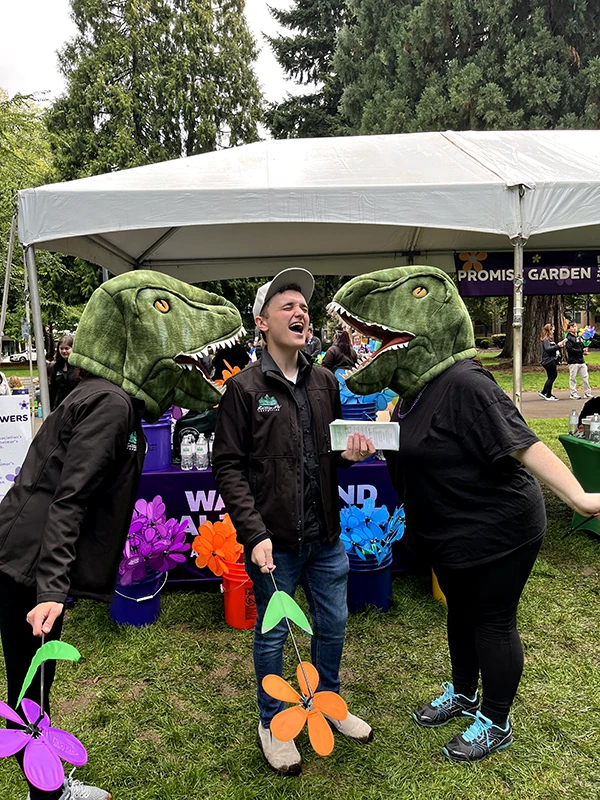 Two people wearing plush dinosaur heads roar at a Heritage employee standing in front of the promise garden flower booth