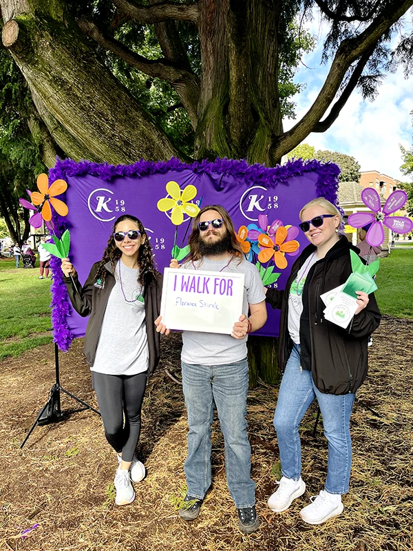 Heritage employees stand in front of a purple banner holding flowers and sign saying I walk for Florence Stirek
