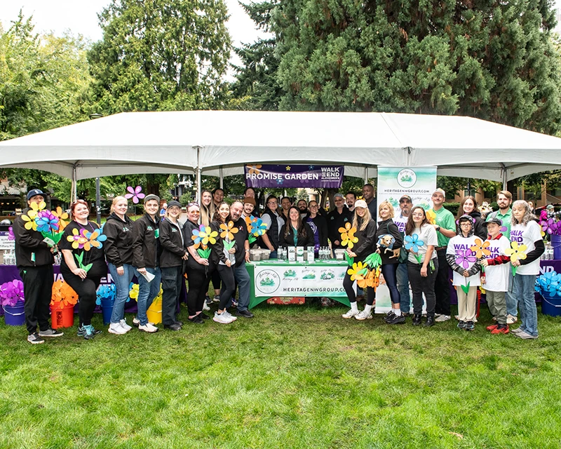 The large group of Heritage employees and family gathers in front of the Promise Garden booth holding flowers