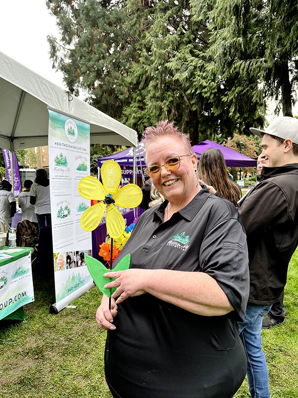 Mona holds a yellow flower and smiles in front of the Heritage booth