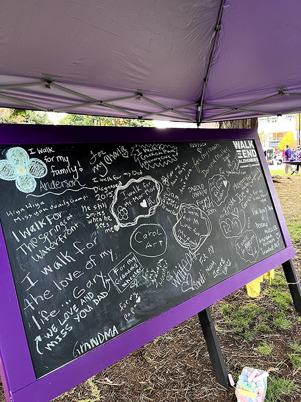 A wide chalkboard with purple border has words in many different people's handwriting stating their reasons for walking to end Alzheimer's