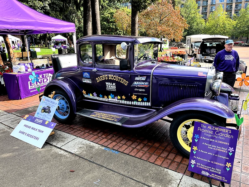 Purple vintage Model A vehicle has stickers with Barb's Boosters, other business logos, Walk to End Alzheimer's logo, and flowers.