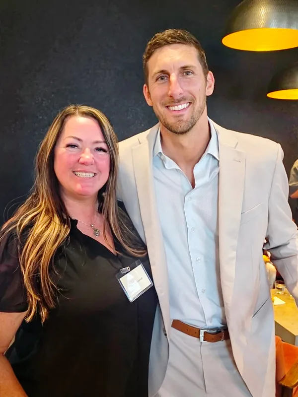 Tara, wearing a black shirt with a nametag clipped to it stands smiling with a man wearing a light blue shirt and tan jacket and pants.
