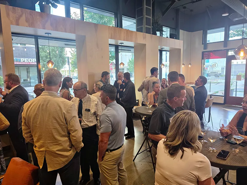 A group of people are standing gathered in small groups of 3-5 people at the Amavi Cellars and Pepper Bridge winery around tall round tables. They are all having conversation and some have wine glasses.